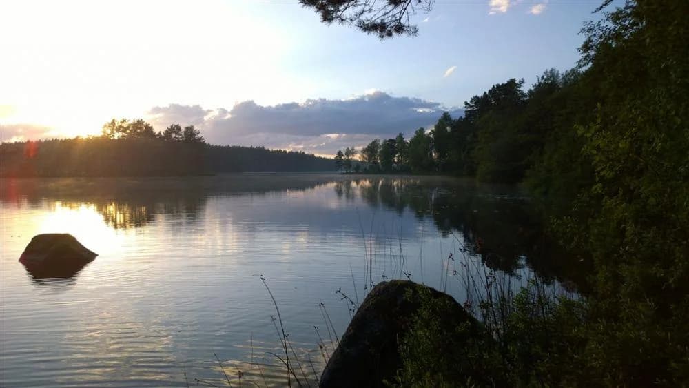 En naturskön sjö med klart blått vatten omgiven av grönska, där himlen speglas i vattenytan. Perfekt för avkoppling och naturupplevelser på Långasjönäs Camping & Stugby.