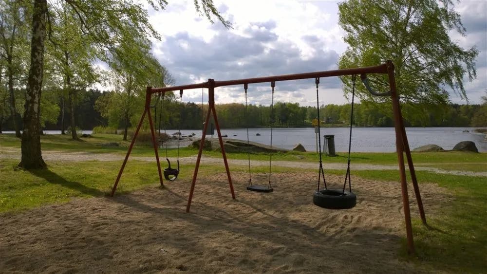 En lekplats i en park med gungor och rutschkana under skuggiga träd, belägen på Långasjönäs Camping & Stugby.