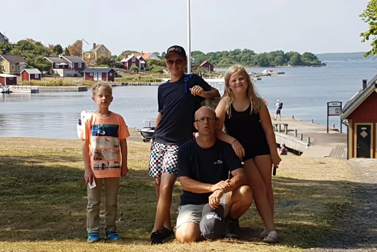 En glad familj njuter av en solig dag vid vattnet på Långasjönäs Camping & Stugby. Barnet bär solglasögon och leker bredvid en liten båt, vilket förstärker känslan av sommarsemester och avkoppling.