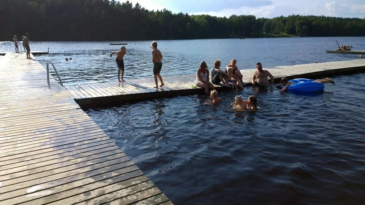 En solig dag vid Långasjönäs camping & stugby; en sjö med en brygga där människor badar och njuter av fritidsaktiviteter vid vattnet.