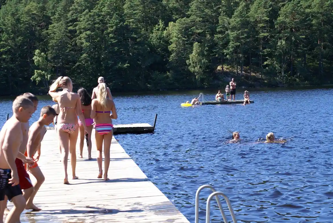 En solig sommardag vid Långasjönäs camping & stugby, där barn och vuxna njuter av lek och bad i en klar sjö. Människor i baddräkter kopplar av vid vattnet och deltar i olika utomhusaktiviteter.