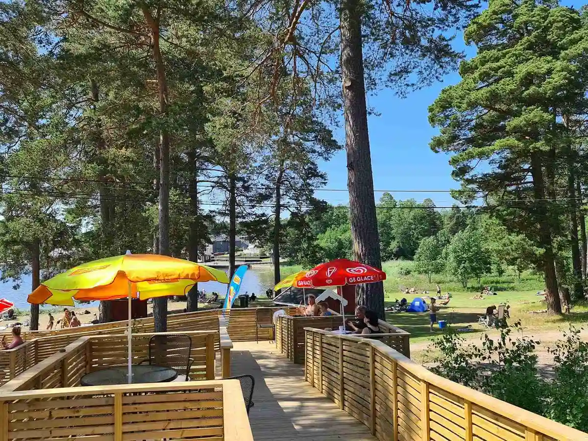En solig dag på Gävle Camping Engesberg med grönskande träd som skapar skugga över picknickbord och en gångstig genom parken. Perfekt för avkoppling och sommaraktiviteter.