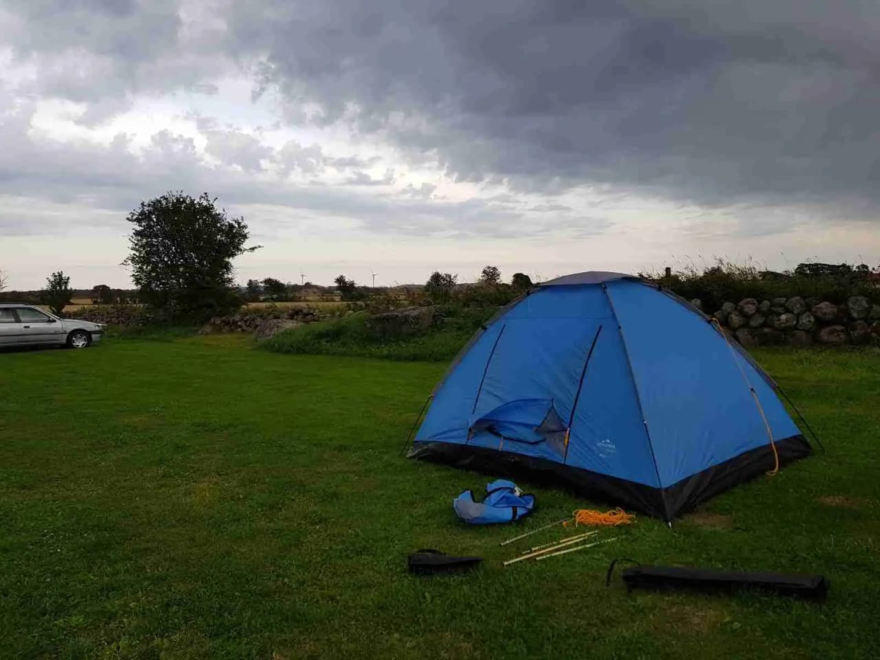 En bild av ett tält uppsatt på Rödlix Hostel & Camping, omgiven av campingutrustning och en prydlig grön park. Tältets triangulära form och tarpaulin kan ses tydligt, vilket ger en känsla av friluftsliv och rekreation.
