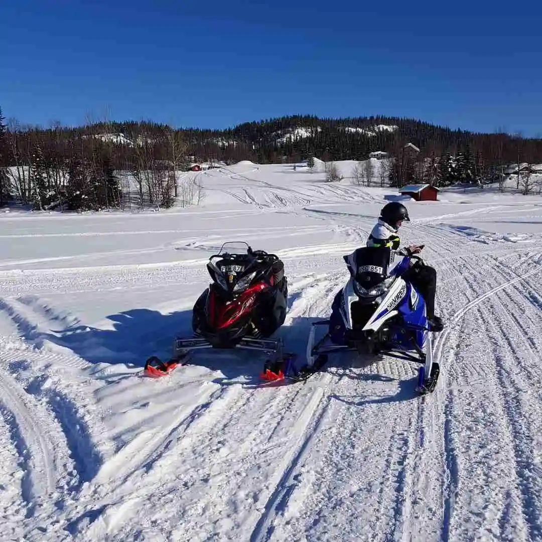 En vintermiljö med snötäckt landskap där en person iklädd vinterkläder och hjälm kör snöskoter i Grönfjälls fritidsby, omgiven av snötäckta kullar och träd.