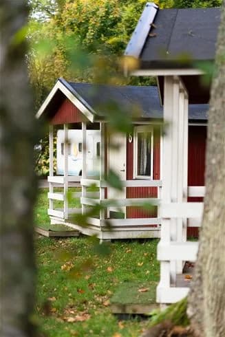 En grönskande campingplats med en stuga omgiven av trädgård och gräsmatta. Träd ger skugga över området som inkluderar en anlagd utomhusstruktur i form av en timmerstuga.