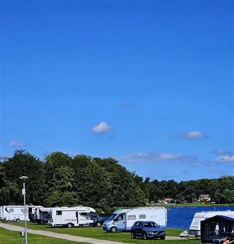 En husvagn står parkerad på en campingplats under en molnig himmel, omgiven av fler campingfordon och husbilar samt lite campingutrustning.