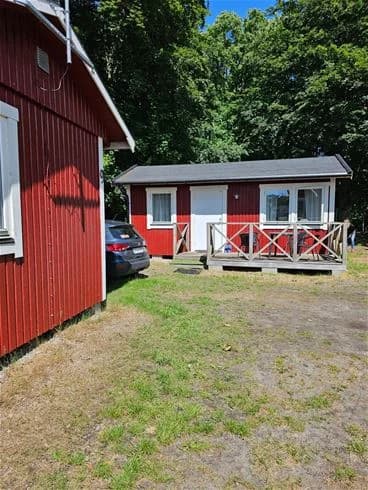 Bild av en campingstuga omgiven av en grönskande trädgård med träd och buskar i bakgrunden. Stugan har enkla träväggar och befinner sig i en skuggig del av trädgården.