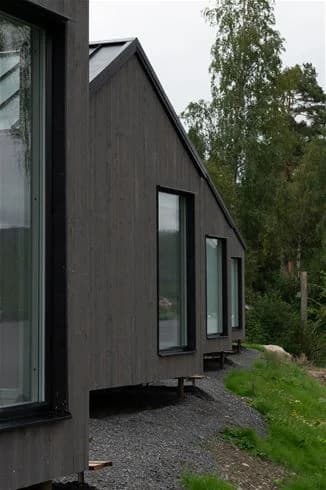 En idyllisk stuga vid Skuleberget Havscamping. Omgiven av gröna växter och träd, med ett bergigt utsprång i bakgrunden och en välskött fasad.