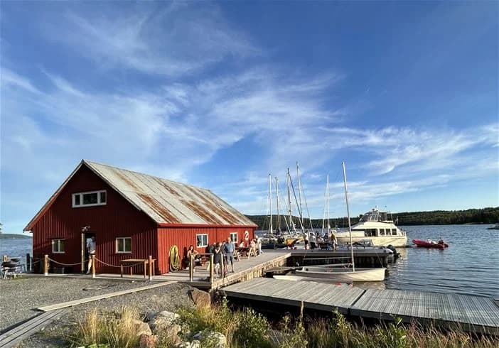 En panoramavy över Skuleberget Havscamping med flera båtar förtöjda vid en träbrygga i småbåtshamnen. Vattnet ligger spegelblankt och omgivningen består av frodig grönska. Masten på en av båtarna sträcker sig mot den klara himlen.