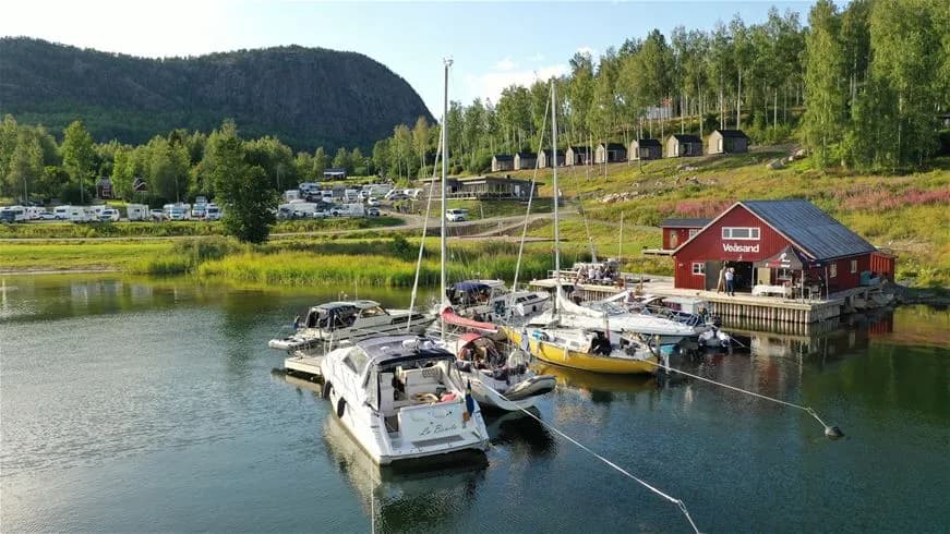 En vy över Skuleberget havscamping med båtar förtöjda vid bryggan, omgivna av glittrande vatten och omgivande berg.