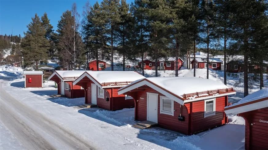 En vinterbild av en timmerstuga vid Skellefteå camping, täckt av snö, med sluttande tak och omgivande vinterlandskap.