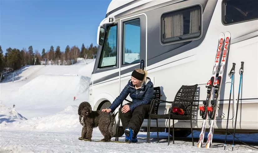 Alt-text: En vinterbild av Skellefteå Camping med snöklädd mark och en husvagn uppställd bland träden.