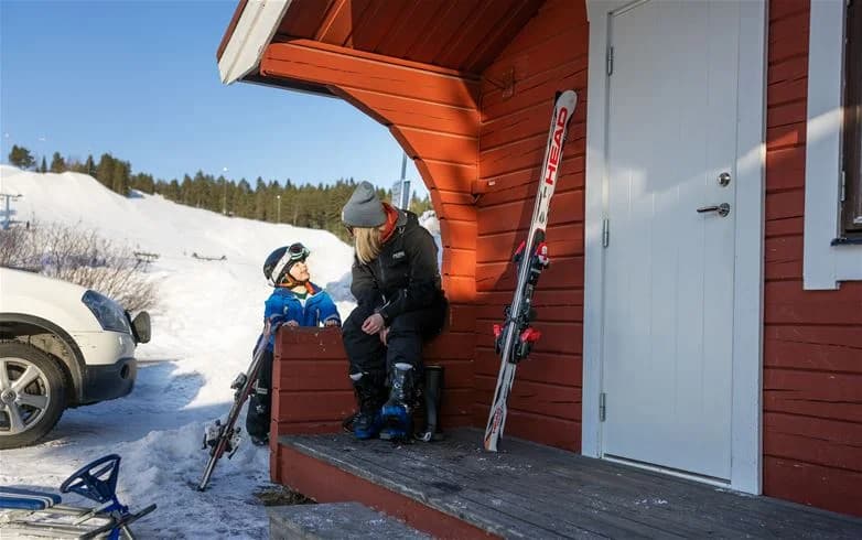 En vintrig landskapsbild av Skellefteå Camping med snötäckta stugor, skidutrustning och i bakgrunden ett snöigt landskap som inbjuder till friluftsaktiviteter.