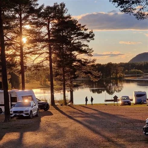 Alt-text: En naturskön kvällsbild vid Sonfjällscampen, där solen sänker sig över en stilla sjö. Färgerna på himlen skiftar i varma nyanser av apelsin och rosa, medan landskapet speglar sig mjukt i det lugna vattnet.