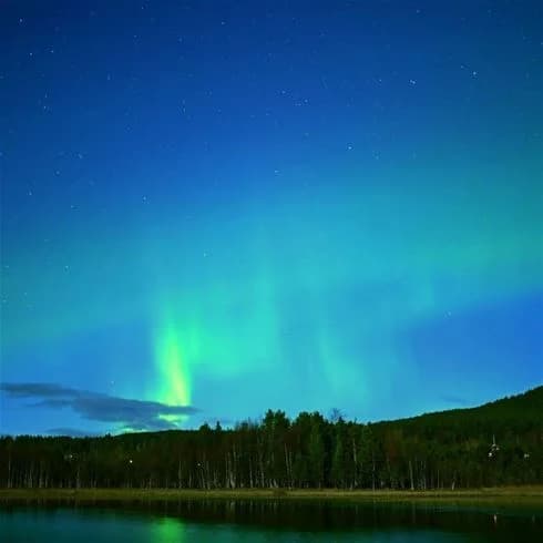 En nattlig vy vid Sonfjällscampen med norrsken som speglas i en stilla sjö omgiven av grön, skogig natur.