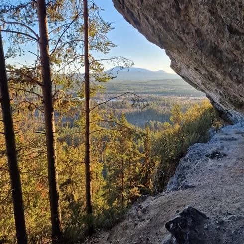 Vy över Sonfjällscampen med storslagen natur, där bergiga landskap och vildmarken dominerar. Skog och stenformationer syns i förgrunden, med mäktiga berg i bakgrunden.