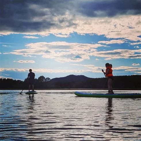 En person som står på en stand-up paddleboard på en lugn sjö omgiven av molntäckt himmel. I bakgrunden syns vidsträckt natur med bergssilhuetter vid horisonten.
