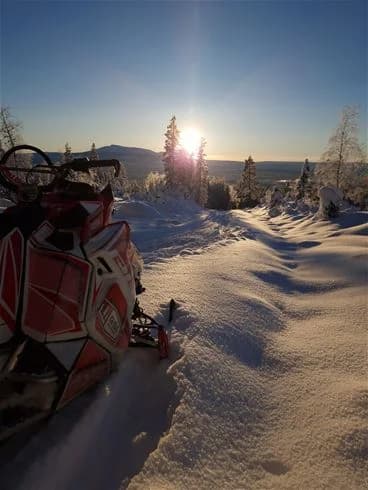En snöskoter står parkerad i ett vinterlandskap täckt av snö och is, omgiven av glaciala formationer. Solen går ner i horisonten, vilket färgar himlen i varma nyanser av orange och rosa. Perfekt för vinteraktiviteter som snöskoteråkning. 