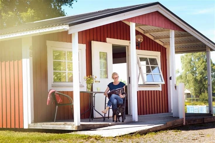 En trästuga med veranda och rött tak omgiven av grönska vid Fyrishov Stugby och Camping.