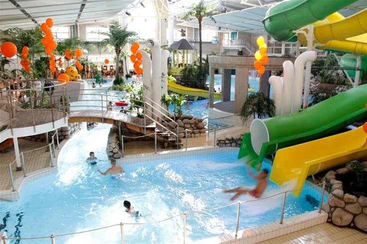 En familj i semestermode vid ett vattenland, barn leker i poolen under färgglada parasoller i Fyrishov Stugby och Camping.