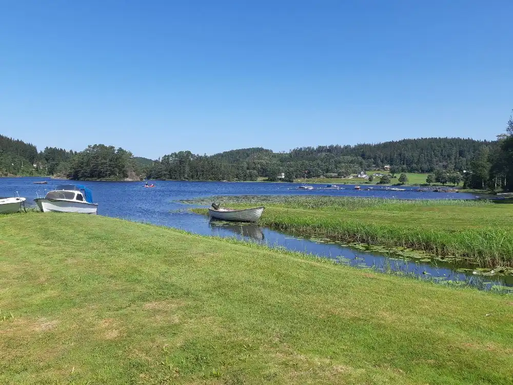 En naturskön vy vid Bullarebygdens familjecamping med en vattenväg som slingrar sig genom ett frodigt landskap av ängar och växtlighet, intill en stilla sjö.