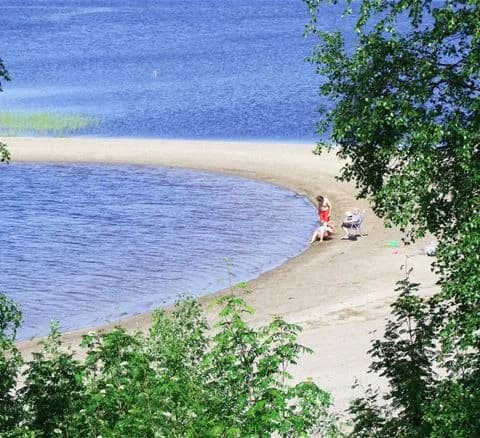 Vy över Särna camping med en glittrande sjö i förgrunden, omgiven av en strand och gröna skogar, som skapar en naturskön plats för semester och rekreation.