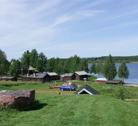 En grön campingplats vid Särna med gräs och ängar, små stugor och skjul omgivna av landsbygdens öppna landskap.