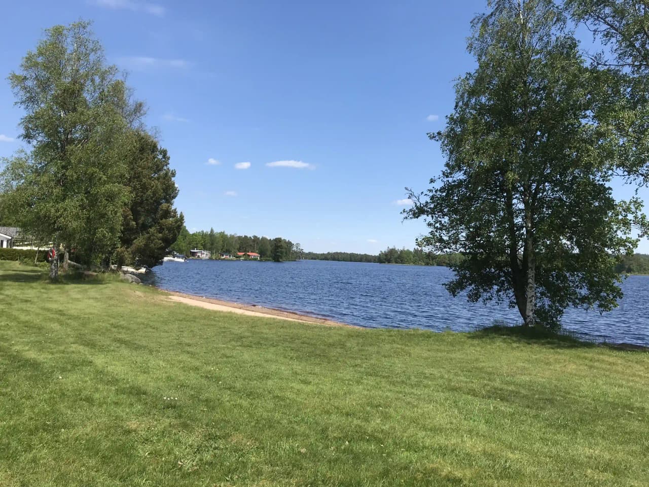 En naturskön vy över Bexet Camping med en stilla blå sjö omgiven av grönska och gräs. Landskapet präglas av det öppna området som gränsar till vattnet, vilket ger en avkopplande atmosfär.