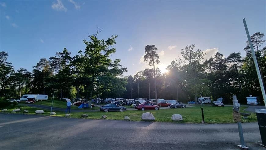 En campingplats med parkering i Trollhättan, omgiven av träd. Flera typer av bilar, inklusive familjebilar och lyxbilar, står parkerade på asfaltsytan.