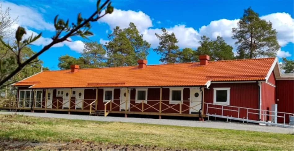 En röd trästuga med veranda omgivet av grönskande gräsmatta och grusvägar på Trollhättans Camping City.