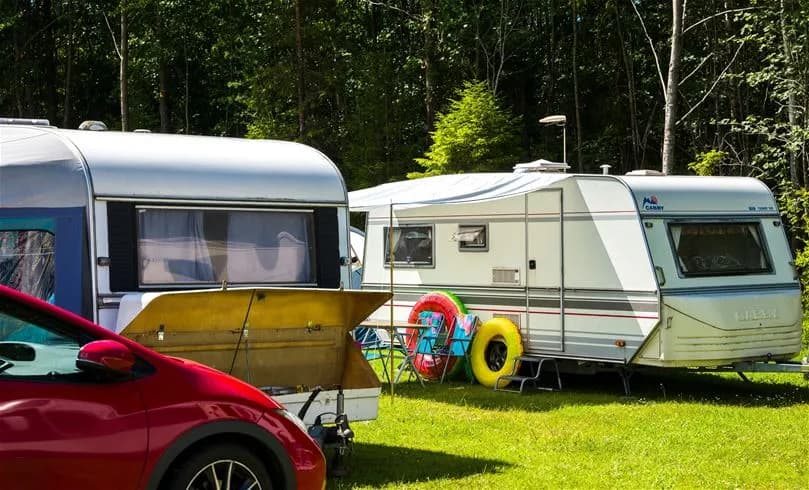 En campingplats med husvagnar och husbilar parkerade på grusplan, omgivna av lummiga träd. Flera tält är uppsatta och picknickbord står utställda, vilket skapar en avkopplande atmosfär.