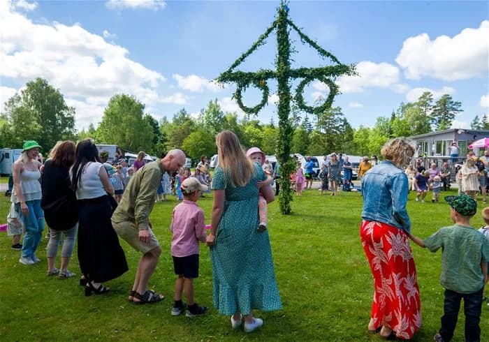 En livlig scen på Stenrösets camping i Trollhättan med en stor samling av människor som njuter av en utomhusaktivitet. Deltagarna ser ut att delta i ett offentligt evenemang som präglas av rekreation och nöje. Vårblommor och grönskande träd omger området, vilket förstärker den gemytliga stämningen.