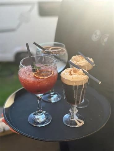 En somrig uteservering på Stenrösets Camping i Trollhättan, med ett bord fyllt av olika rätter och drycker, inklusive en färgglad dessertcocktail med sugrör.