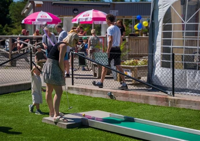 En grupp människor spelar minigolf på en konstgjord gräsmatta vid Stenrösets Camping i Trollhättan. I bakgrunden syns en grönskande park.