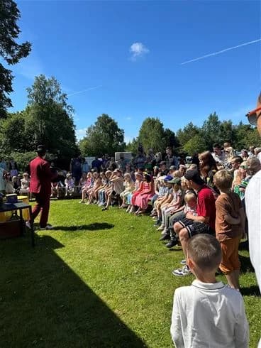 En folkmassa i sommarkläder samlad på Stenrösets camping i Trollhättan, omgiven av grönskande träd. Vissa har t-shirts och skjortor, vilket skapar en avslappnad och social atmosfär.