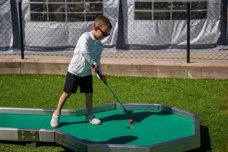Alt-text: "Besökare spelar minigolf på Stenrösets Camping i Trollhättan, omgiven av grönska och campingmiljö."