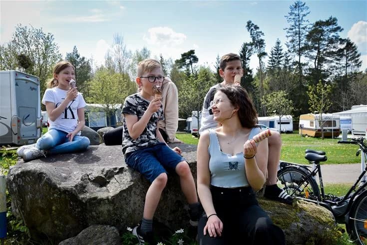 En solig dag vid Stenrösets Camping i Trollhättan, med människor som njuter av utomhusaktiviteter. Några har picknick i parken och andra är aktiva med olika fritidssysselsättningar; en känsla av sommarsemester och avkoppling infinner sig.