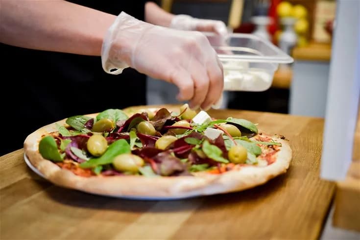 En nygräddad pizza ligger på ett rustikt träbord. Pizzan är toppad med smält ost, skinka, champinjoner och färska örter. I bakgrunden syns några köksredskap.