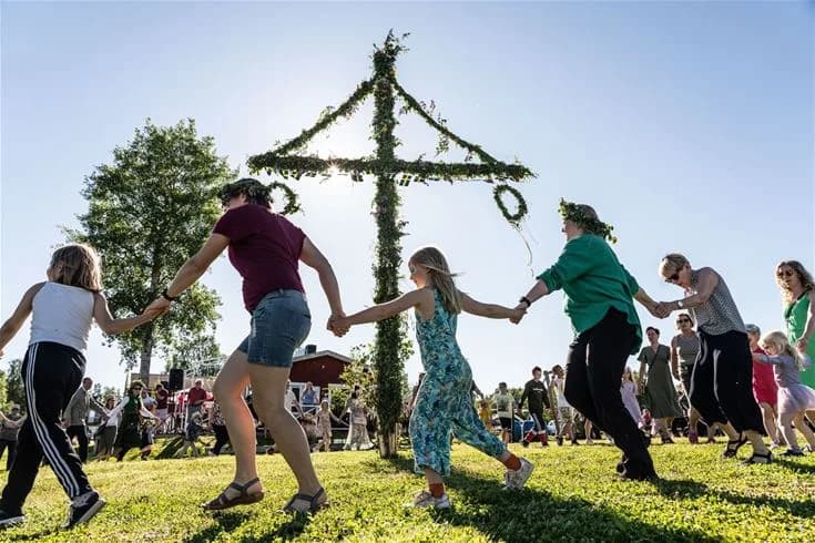 En folkmassa samlas för att fira en traditionell högtid vid Kukkolaforsen Turist & Konferens, där en majstång pryder scenen och festligheterna inkluderar underhållning och kulturella uppträdanden i en naturskön miljö.