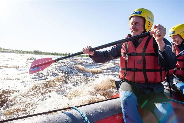 En grupp människor i säkerhetsutrustning paddlar en båt i Kukkolaforsens forsande vatten. De bär hjälmar och flytvästar och deltar i ett spännande vattensportäventyr.