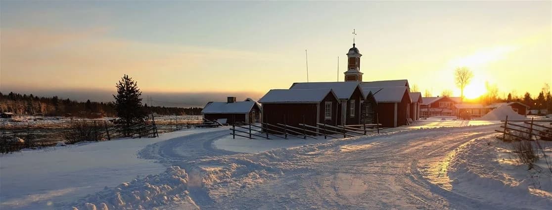 Snöigt vinterlandskap vid Kukkolaforsen Turist & Konferens, med stugor och frostiga träd i skymningen. Snön täcker marken och ger en lugnande vy av den omgivande byn och sluttningen.