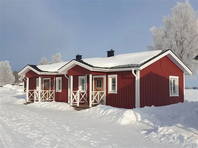 En snötäckt stuga vid Kukkolaforsen under vintern, med ett snöigt tak och ett fruset landskap runt omkring.