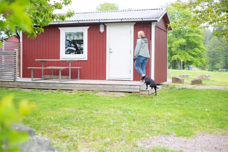 En rödmålad stuga med vita knutar står i grönskande omgivning. Stugan har en dörr och flera fönster, omgiven av välskötta gräsmattor och träd.