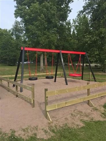 Bilden visar en lekplats med gungor och en rutschkana omgiven av gröna gräsmattor och skuggande träd i en öppen parkmiljö vid Kinnekulle Camping & Stugby.