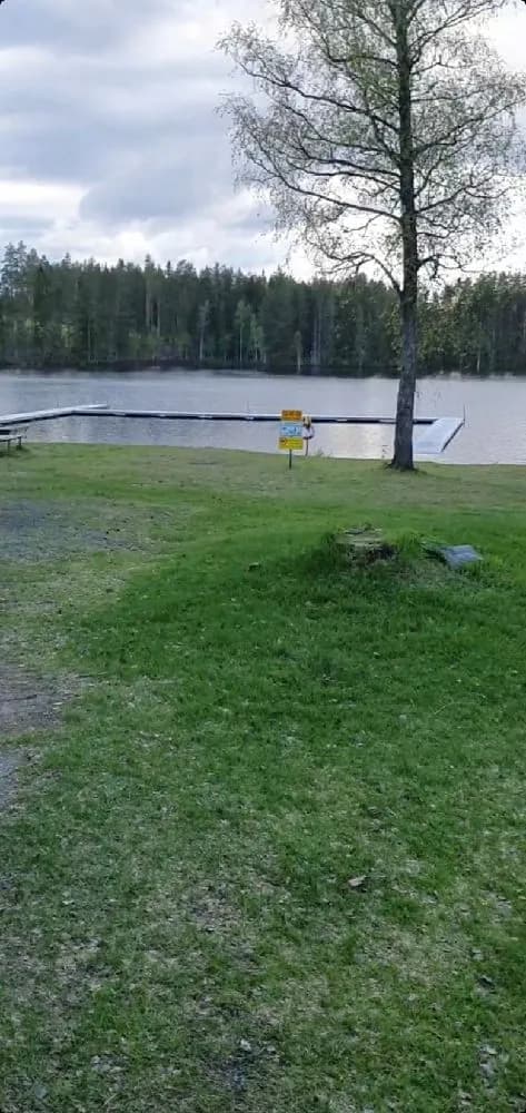 En grönskande campingplats med skuggiga områden, en välskött gräsmatta och en närliggande sjö. Det finns utomhusmöbler och bänkar för avkoppling.
