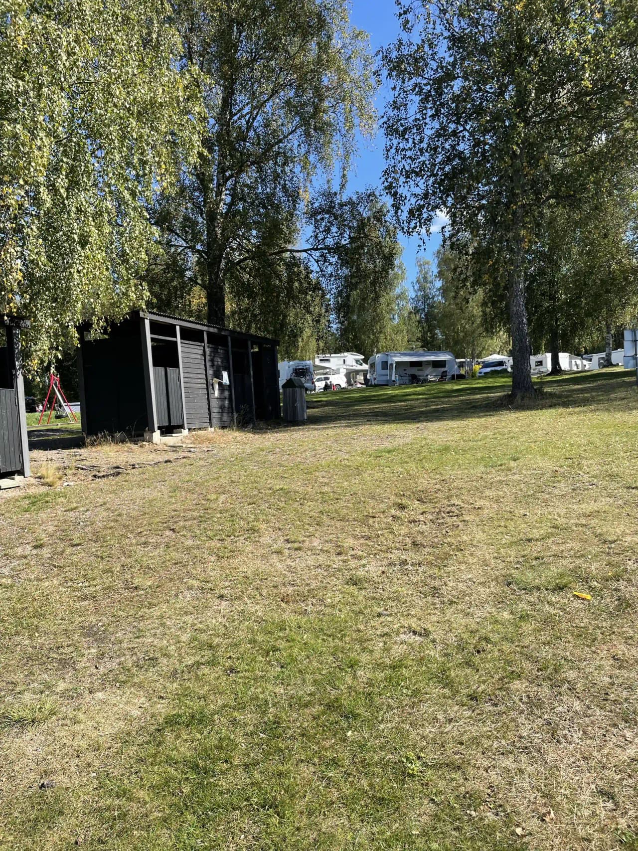 En grönskande campingplats omgiven av träd och skuggiga områden, med parkerade husvagnar och bilar på en gräsbevuxen yta under en klar himmel.