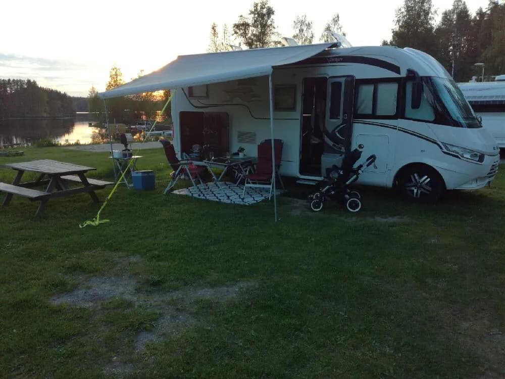 En husvagn med uppfällt förtält står parkerad på en campingplats, omgivet av gröna träd och gräs. En bil och husbil syns i närheten.