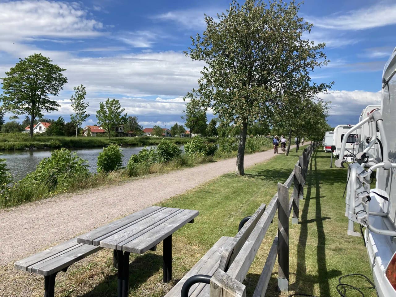En campingplats i Töreboda med parkerade husbilar och husvagnar vid en kanal. En bänk står i parken nära vattnet, omgiven av grönska och våtmarker.