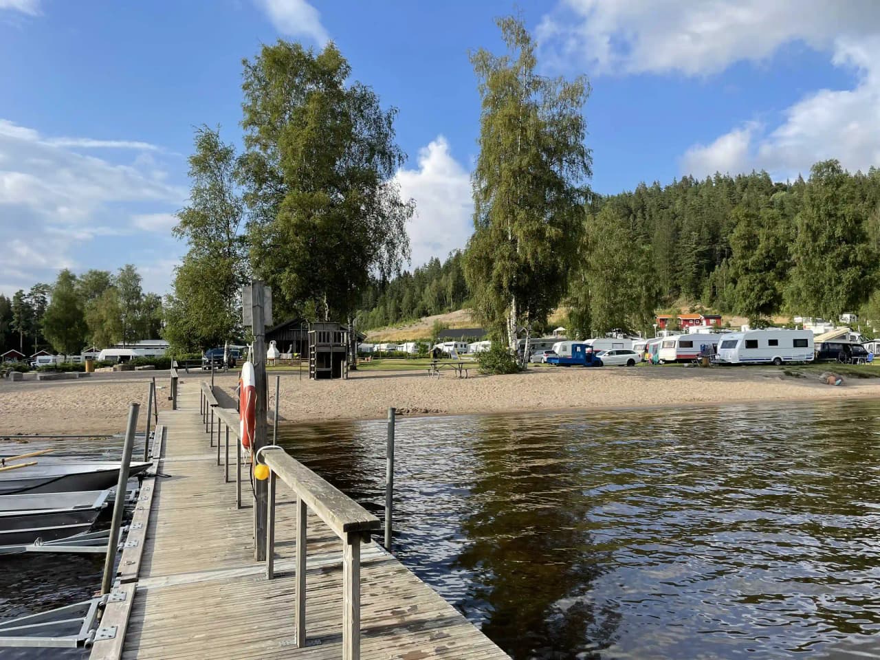 En liten båt ligger vid stranden av Ragnerudssjön, omgiven av gröna kullar och klarblått vatten, med campingområdet synligt i bakgrunden.