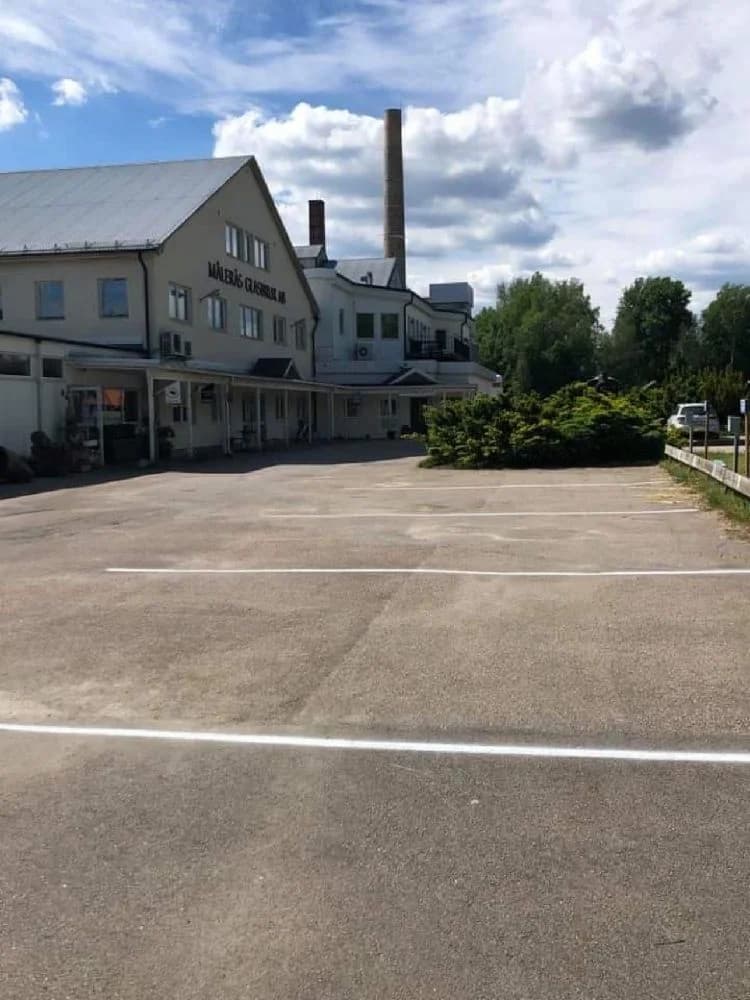 Bild på Målerås Glasbruk med en parkeringsplats i förgrunden och en värdshusliknande byggnad i bakgrunden.